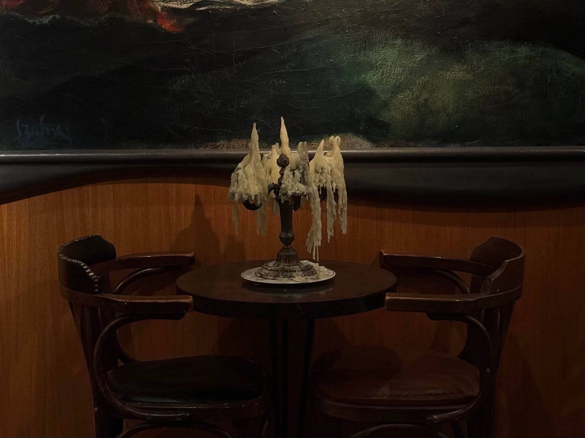 A candelabra heavy with drips of old wax on a small round café table between two bentwood armchairs, beneath a dark painting.