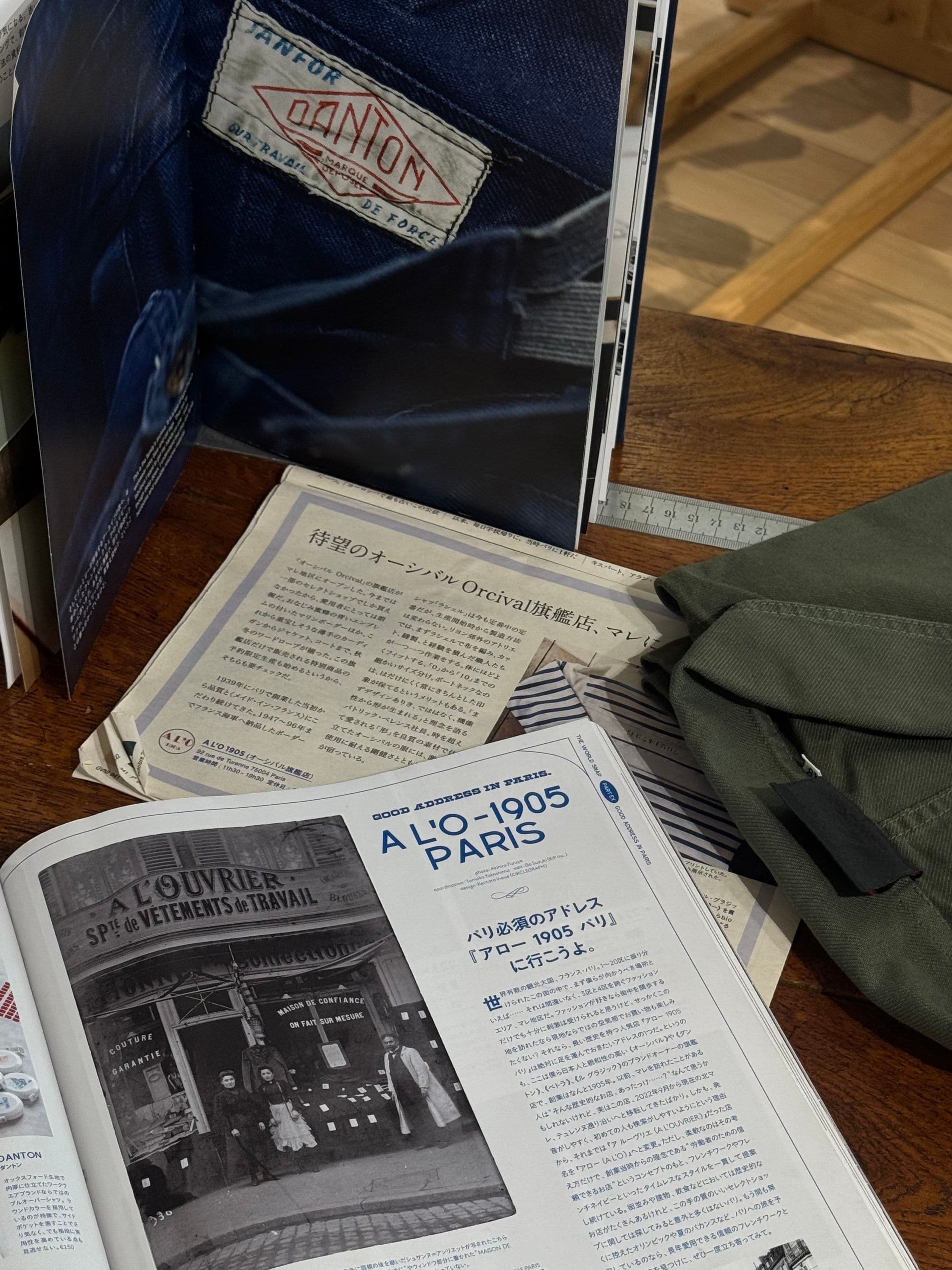 Open magazines and a Japanese newspaper clipping about French workwear spread across a wooden table beside a folded olive jacket.