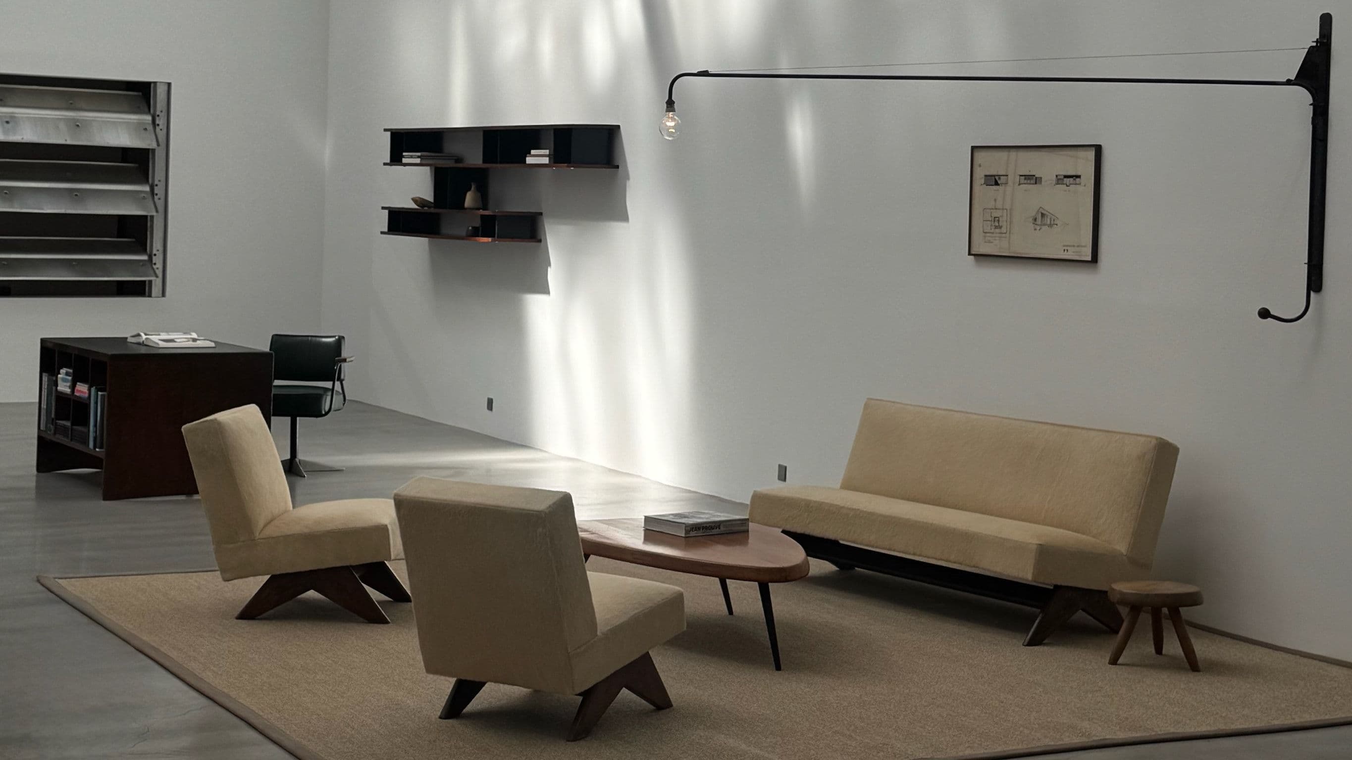 A pale gallery room with two upholstered lounge chairs and a sofa on a sisal rug around a low wooden table, beneath a long wall-mounted swing-arm lamp.