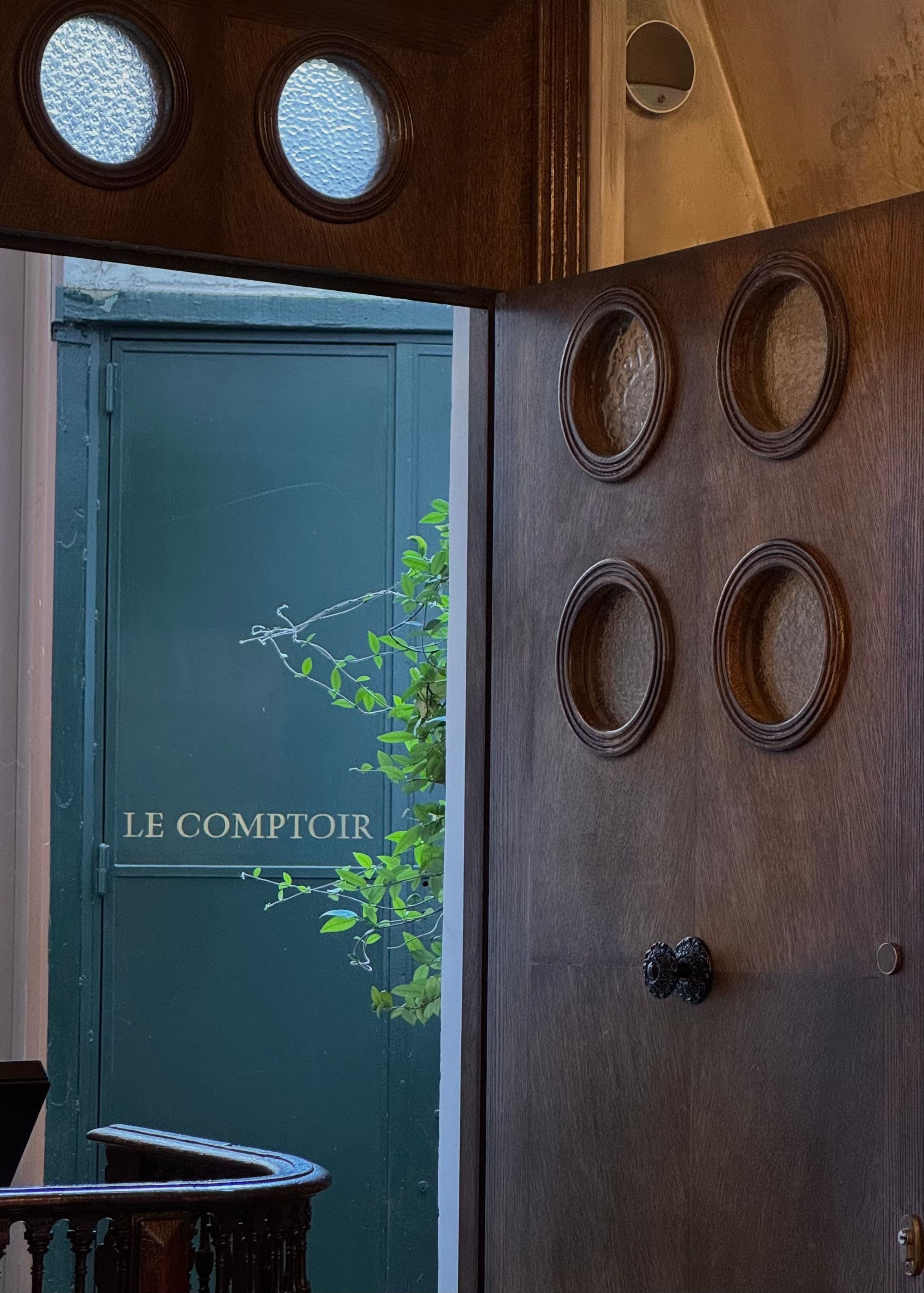 An open wooden door set with round porthole windows, looking through to a green door lettered “Le Comptoir” and a climbing plant.
