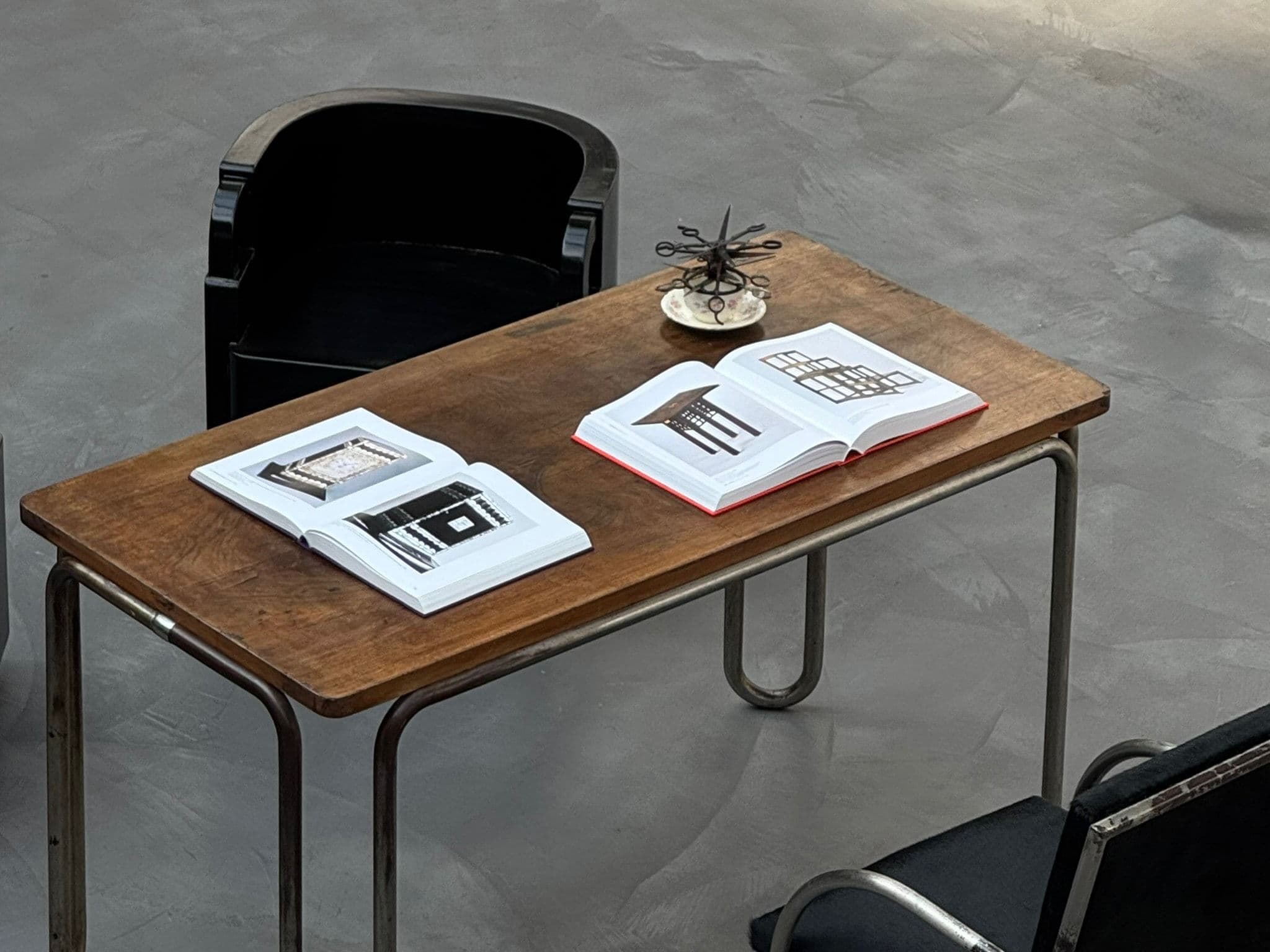 Two open books on a worn wooden desk with tubular steel legs, seen from above beside a black chair.